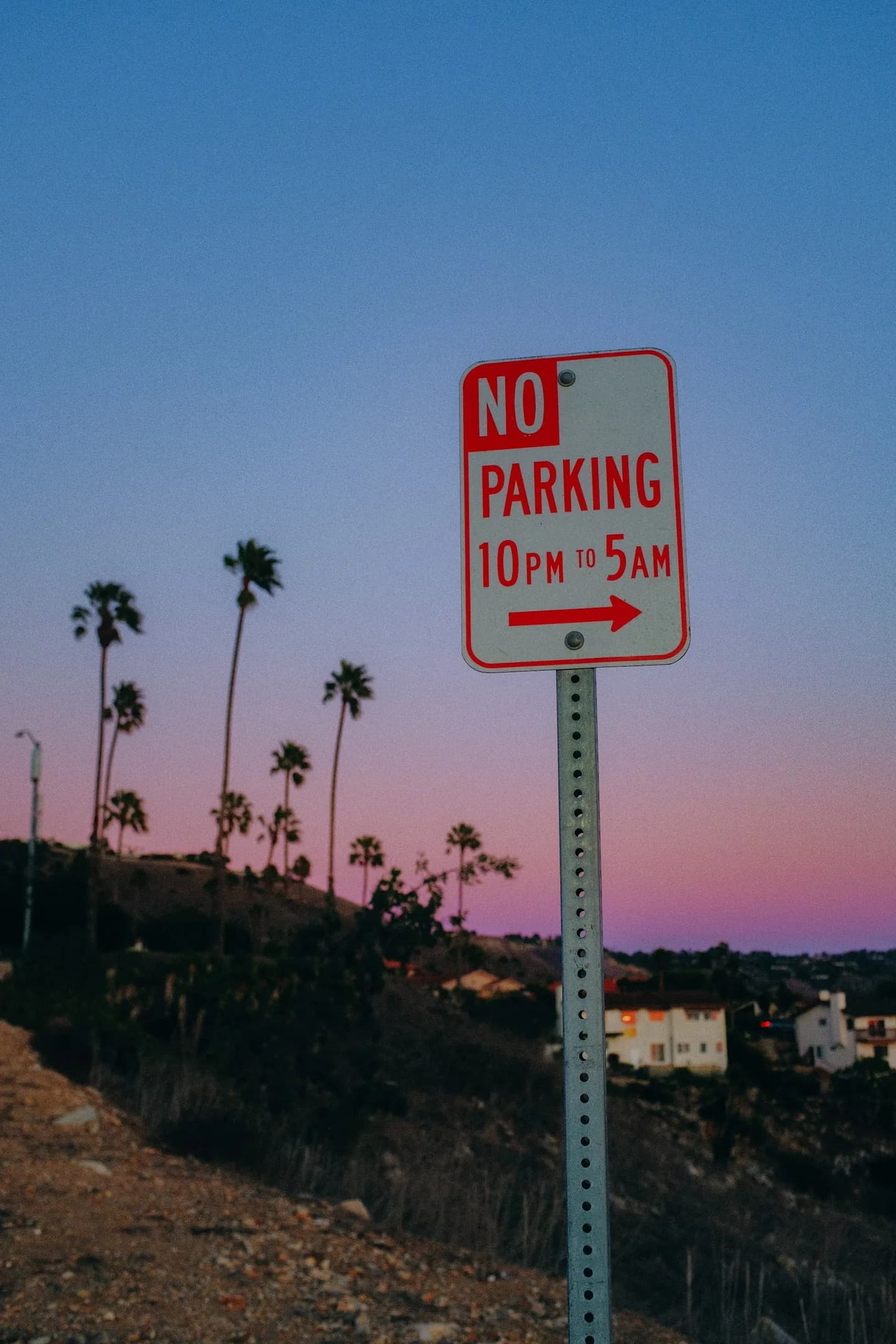 A roadside sign with hillside homes and palms beneath a pastel sky